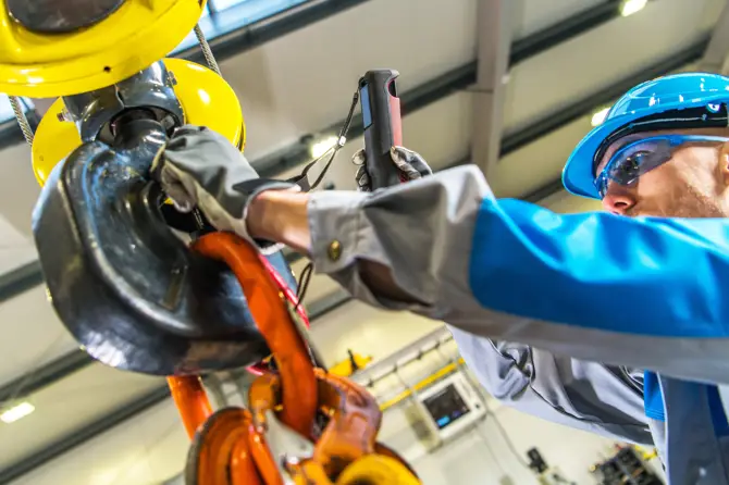 Inspector wearing a blue jumpsuit and PPE performing lifting equipment inspections on a large industrial hook. Inspector wearing a blue jumpsuit and PPE performing lifting equipment inspections on a large industrial hook.